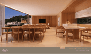 Área interna do salão de festas decorada com móveis de madeira clara e tons neutros. O espaço conta com mesas de jantar, bancada gourmet de mármore e uma grande abertura para a área externa do terraço.